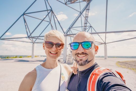 Ein lächelndes Paar mit Sonnenbrille macht ein Selfie im Freien vor einer großen, geometrischen Metallstruktur unter einem sonnigen, blauen Himmel - und hält damit ihr Abenteuer "Tief im Westen" fest. Die Frau hat kurzes blondes Haar und der Mann hat einen rasierten Kopf und Bart.