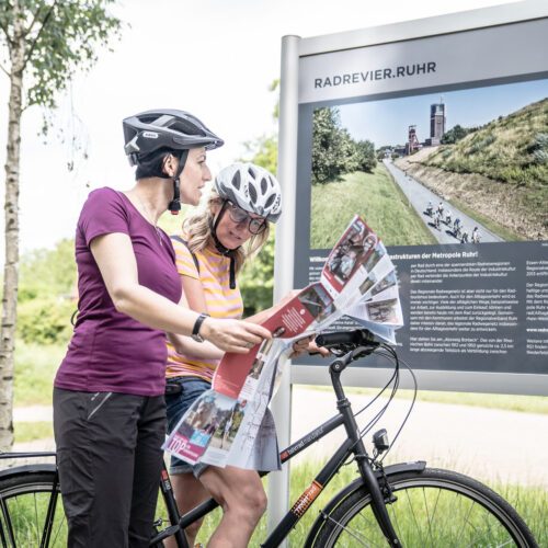 Das Foto zeigt zwei Radfahrerinnen mit Übersichtskarte vor einer radrevier Infotafel auf der Rheinischen Bahn in Essen