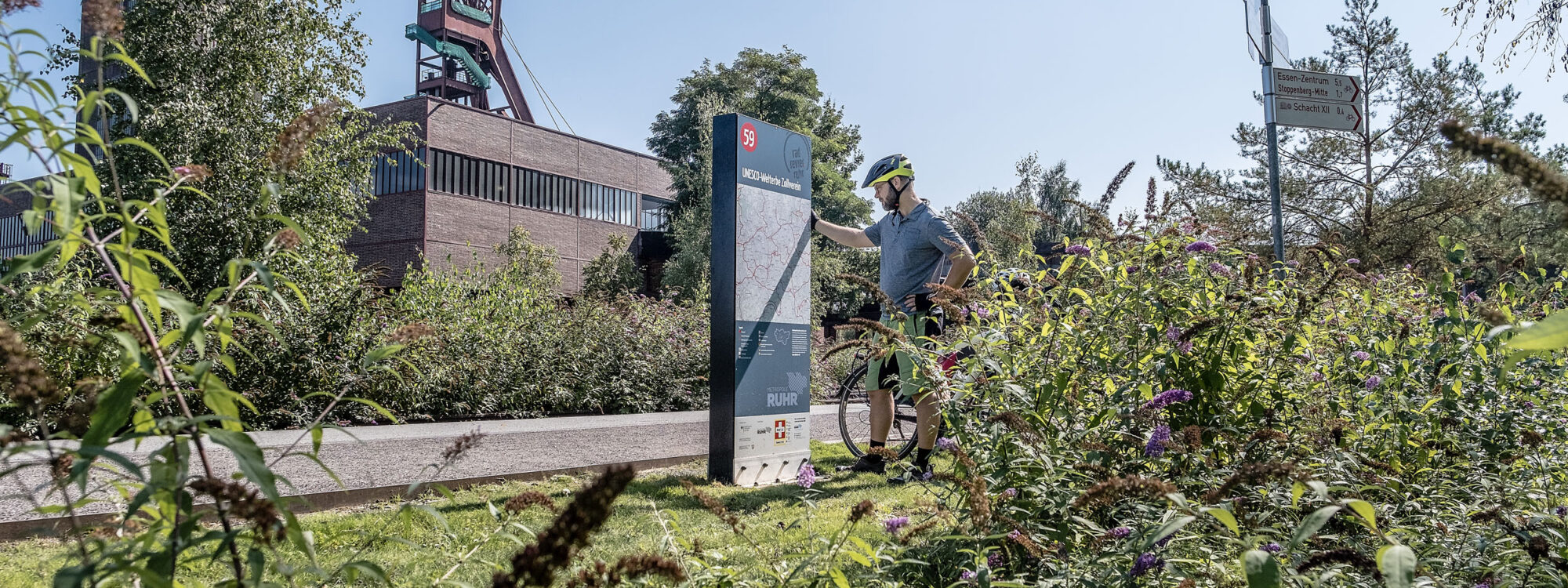 Das Foto zeigt einen Radfahrer auf dem Gelände des UNESCO-Welterbe Zollverein in Essen an der Knotenpunkttafel des Knotenpunkts 59 auf der RevierRoute Grubenfahrt