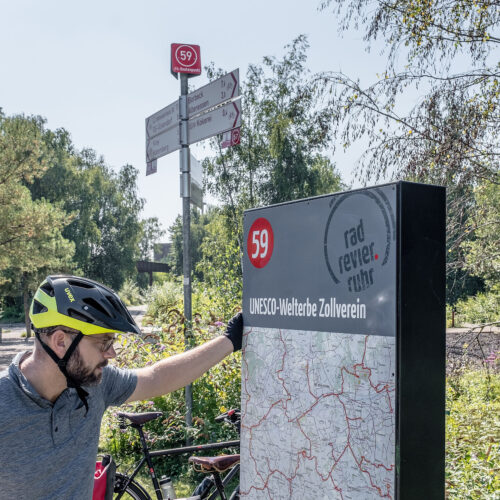 Das Foto zeigt einen Radfahrer auf dem Gelände des UNESCO-Welterbe Zollverein in Essen an der Knotenpunkttafel des Knotenpunkts 59 auf der RevierRoute Grubenfahrt