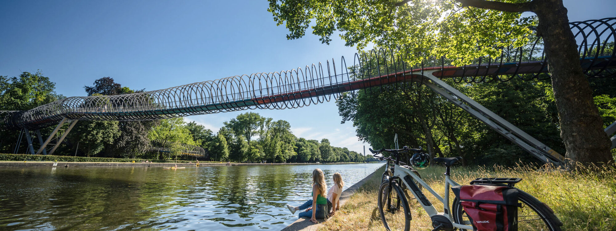 De foto toont twee fietsers aan de oevers van het Rhein-Herne-kanaal voor de Slinky Springs to Fame-brug in Oberhausen