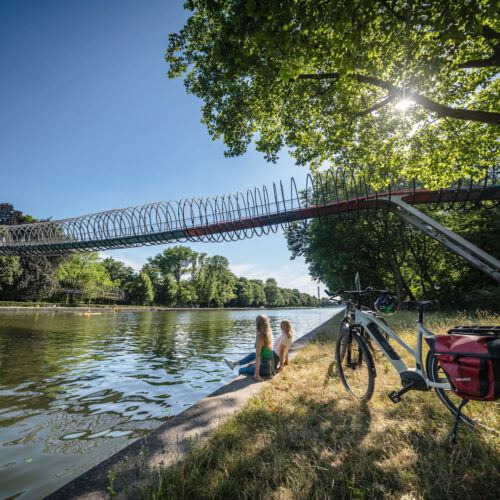 Das Foto zeigt zwei Radfahrerinnen am Ufer des Rhein-Herne-Kanals vor der Brücke Slinky Springs to Fame in Oberhausen