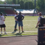 Eine Gruppe von fünf Personen, darunter ein Kameramann, steht auf einem Sportplatz in der Nähe eines Stadions und unterhält sich. Das Wetter ist sonnig, und im Hintergrund sind Bäume und leere Tribünen zu sehen.