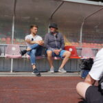 Zwei Männer sitzen und unterhalten sich auf roten Stadionstühlen; der eine trägt ein weißes Hemd und Jeans, der andere eine graue Jacke, Shorts und einen Eimerhut. Eine dritte Person in einem weißen Hemd mit Kopfhörern filmt sie aus dem Vordergrund.