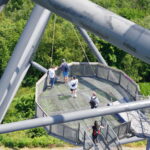 Man steht und geht auf einer erhöhten, geometrischen Aussichtsplattform aus Metall, die von üppigen grünen Bäumen umgeben ist und von großen Metallträgern eingerahmt wird.