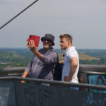 Zwei Männer stehen auf einer Plattform mit Graffiti im Freien, der eine in grauem Hemd und Mütze, der andere in weißem Hemd, der ein Selfie mit einem roten Tablet macht und dabei zusieht. Im Hintergrund breitet sich eine malerische Landschaft unter einem klaren Himmel aus.