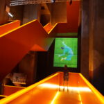 Eine Person steht auf einer orange beleuchteten Rampe in einem modernen Gebäude und blickt auf einen großen grünen Bildschirm, auf dem das Bild eines Fußballspielers in der Mitte des Fußballs an der Wand davor zu sehen ist.