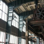 Große Industriefenster lassen Sonnenlicht in das Innere einer Fabrik mit freiliegenden Balken, Metallstrukturen und großen Zahnrädern über den Köpfen und schaffen eine helle und mechanische Atmosphäre.