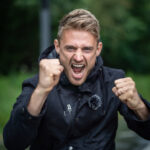 Ein junger Mann in einer schwarzen Jacke steht im Freien, hebt beide Fäuste und schreit mit entschlossener Miene. Hinter ihm sind grüne Bäume und ein verschwommener Hintergrund zu sehen.