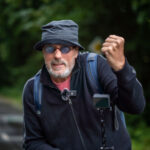Ein Mann, der einen Eimerhut, eine Sonnenbrille und eine schwarze Jacke trägt, steht mit einem Fahrrad im Freien. Er hat einen Rucksack, ein Mikrofon an seiner Jacke befestigt und eine Kamera auf dem Fahrrad montiert und hebt eine Hand zu einer Geste. Im Hintergrund ist üppiges Grün zu sehen.