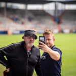 Zwei Männer stehen auf einem Sportplatz. Der eine macht ein Selfie mit einem Smartphone, während der andere daneben posiert. Hinter ihnen sind Stadionplätze und ein unscharfer Hintergrund zu sehen.