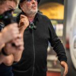 Ein Mann mit einem schwarzen Hut, einer blauen Brille, einer schwarzen Jacke mit Reißverschluss und einer orangefarbenen Hose steht in einem Haus und gestikuliert mit seinen Händen. Eine weitere Person mit Ausrüstung ist teilweise neben ihm zu sehen.