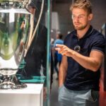 Ein Mann in einem marineblauen Polohemd und grauen Shorts steht vor einer großen Trophäe, die in einer Glasvitrine ausgestellt ist, und deutet mit der rechten Hand auf sie. Die Trophäe hat zwei große Griffe und eine spiegelnde Oberfläche.