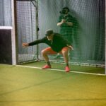 Eine Person, die eine Mütze, ein schwarzes Hemd und rote Shorts trägt, hockt vor einem Tor auf einem Hallenfußballfeld und bereitet sich darauf vor, einen Schuss zu blocken, während eine andere Person hinter dem Netz steht.