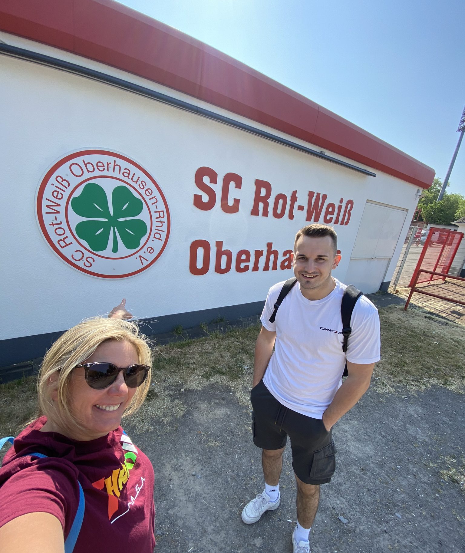 Eine Frau und ein Mann stehen lächelnd vor einem Gebäude mit einem Schild mit der Aufschrift SC Rot-Weiß Oberhausen und einem großen grünen Kleeblatt-Logo. Die Frau zeigt auf das Schild, beide tragen Freizeitkleidung mit Rucksäcken.