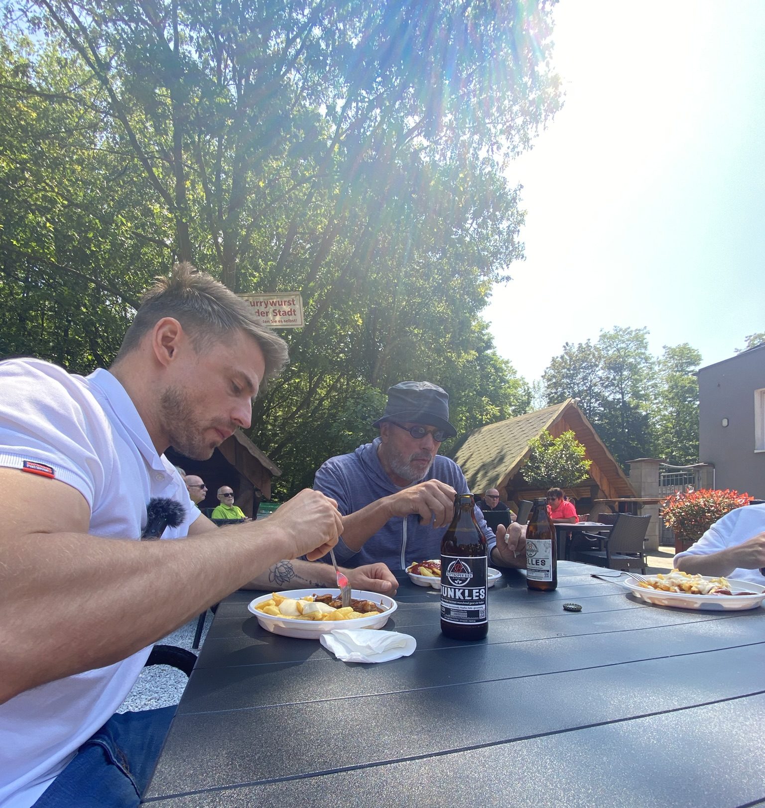 Zwei Männer sitzen an einem sonnigen Tag an einem Tisch im Freien und essen. Einer hat ein weißes Hemd an, der andere trägt eine Sonnenbrille und einen Hut. Eine Bierflasche und Teller mit Essen stehen vor ihnen. Im Hintergrund sind Bäume und kleine Gebäude zu sehen.