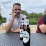 Ein lächelnder Mann in einem weißen Hemd sitzt an einem Tisch im Freien und hat zwei Flaschen Lauder-Bier vor sich stehen. Eine weitere Person ist teilweise rechts zu sehen. Im Hintergrund sind Bäume und ein Plastikstuhl zu sehen.