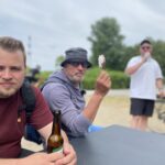 Drei Männer sitzen im Freien an einem Tisch. Ein Mann in einem roten Hemd sitzt mit einer Bierflasche in der Hand, ein anderer mit Hut und Sonnenbrille hält ein Eis in der Hand, und ein dritter Mann in einem weißen Hemd steht im Hintergrund und trinkt.