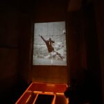 Ein Schwarz-Weiß-Foto eines Fußballtorwarts, der einen Ball abwehrt, wird auf eine hohe Wand in einem schwach beleuchteten Raum projiziert, während orangefarbene Lichter die Bänke unter der Projektion beleuchten.
