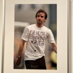 Ein Mann, der ein weißes T-Shirt mit der Aufschrift FRIEDEN STATT KRIEG und einem Schießscheiben-Symbol trägt, steht im Freien und ist auf einem gerahmten Foto zu sehen, das an einer Wand hängt.