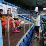 Zwei Männer sitzen auf einem Sofa auf einem Podest in einem Sportstadion; einer spielt Gitarre. Zwei andere, einer mit einer Kamera und einer mit einem Mikrofon, filmen sie. Gerahmte Bilder hängen an einer Wand hinter dem Sofa. Blaue Stadionsitze sind zu sehen.