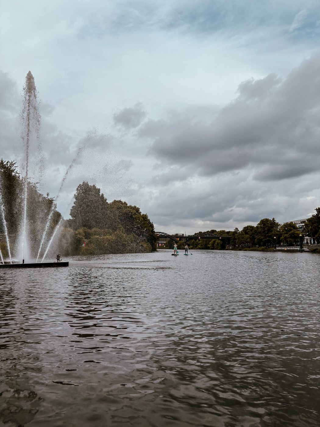 Ein großer Teich mit einer hohen Wasserfontäne auf der linken Seite, umgeben von Bäumen und bewölktem Himmel. In der Ferne sind zwei Personen beim Paddeln zu sehen.