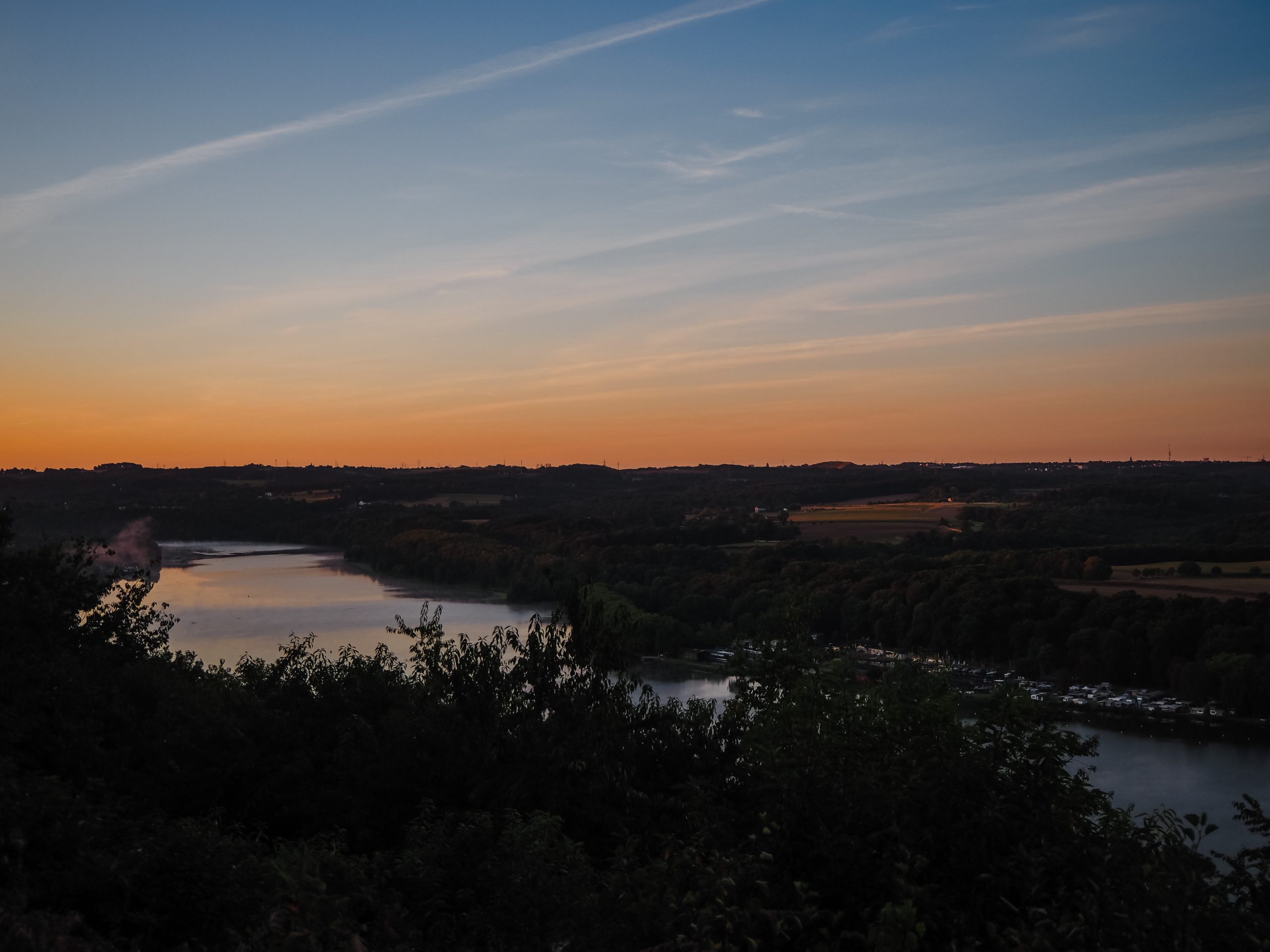 Ein ruhiger Fluss schlängelt sich durch eine üppige, grüne Landschaft bei Sonnenuntergang, mit orangefarbenen und blauen Farbtönen am Himmel und Silhouetten von Bäumen und Hügeln in der Ferne.