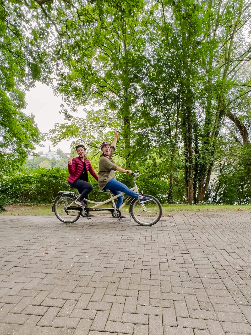 Zwei Personen mit Helmen fahren auf einem Tandemfahrrad auf einem gepflasterten Weg, der von üppigen grünen Bäumen umgeben ist. Sie lächeln und heben jeweils eine Hand zu einer fröhlichen Geste.