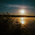 Sonnenaufgang über einem ruhigen See mit aufsteigendem Nebel, silhouettierten Pflanzen und Bäumen im Vordergrund und einem klaren Himmel mit ein paar Kondensstreifen.