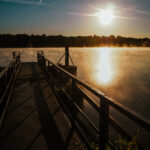 Ein Metallsteg mit Geländer führt über einen ruhigen See bei Sonnenaufgang, wenn Nebel vom Wasser aufsteigt und die Sonne über dem baumbestandenen Horizont scheint. Schatten erstrecken sich über den Steg.