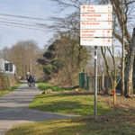 Das Foto zeigt einige Hinweisschilder für Radfahrer auf der Grugatrasse in Essen, auf der es sich fernab des Straßenverkehrs entspannt radeln lässt.